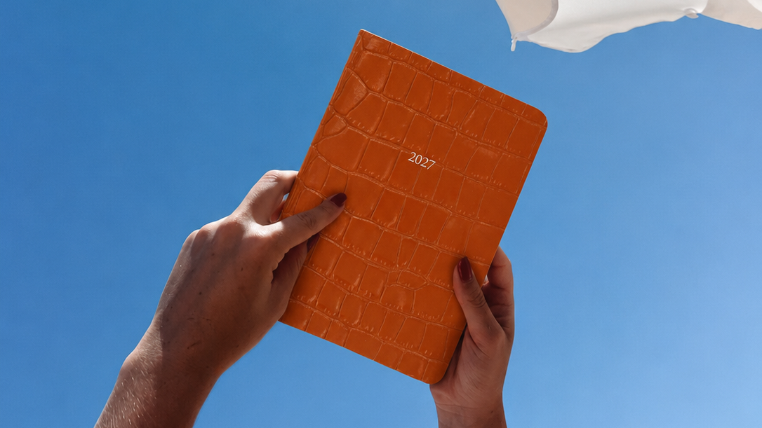 An orange crocodile embossed diary with '2027' printed on the cover, held up by two hands under a white umbrella against a clear blue sky.